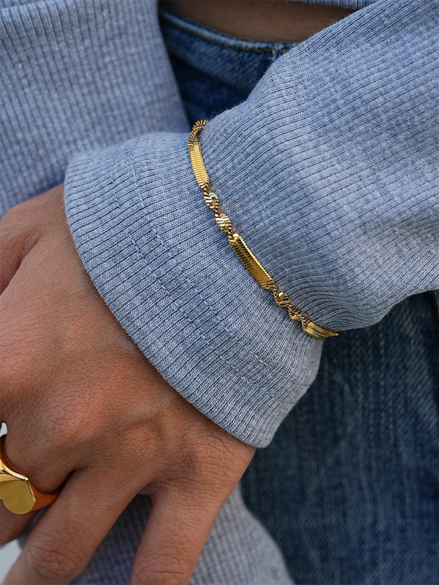 Getragenes goldfarbenes Armband mit flacher, gedrehter Kette am Handgelenk, grauer Rippstrick-Ärmel und Jeansstoff.