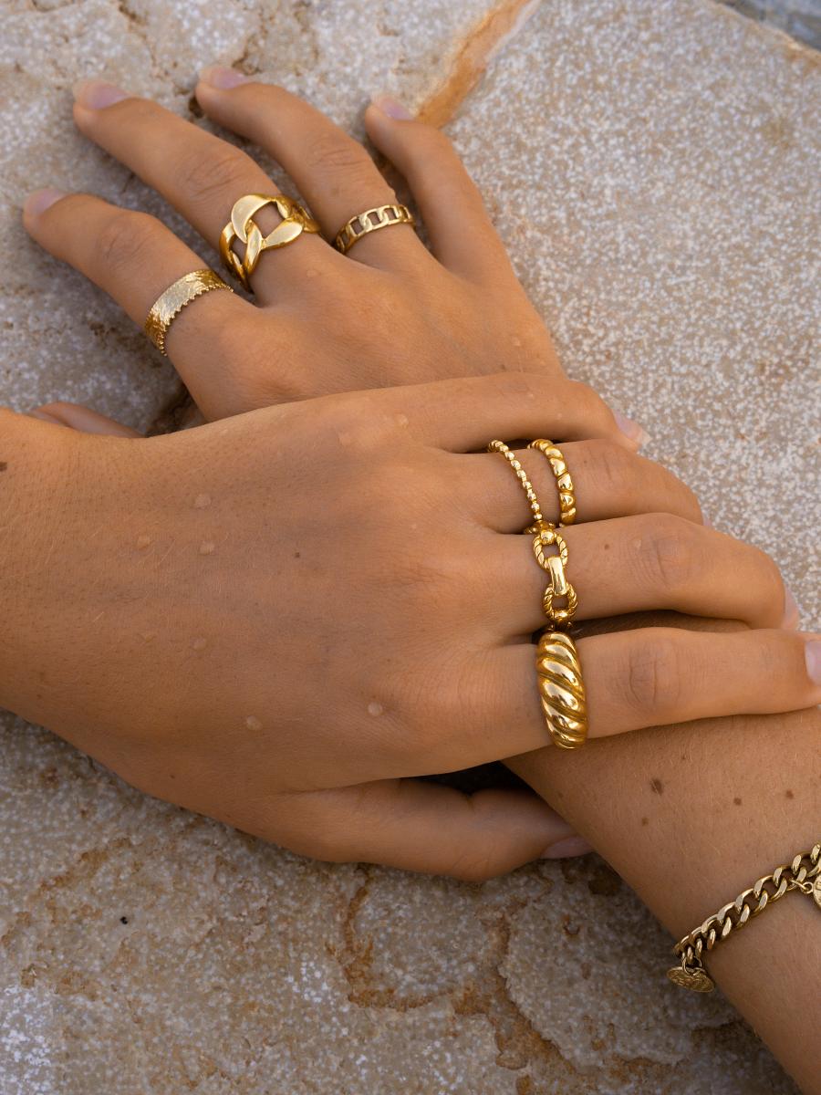 Zwei Hände mit mehreren goldfarbenen Fingerringen auf rauem Stein, darunter gewundene und kugelige Modelle.