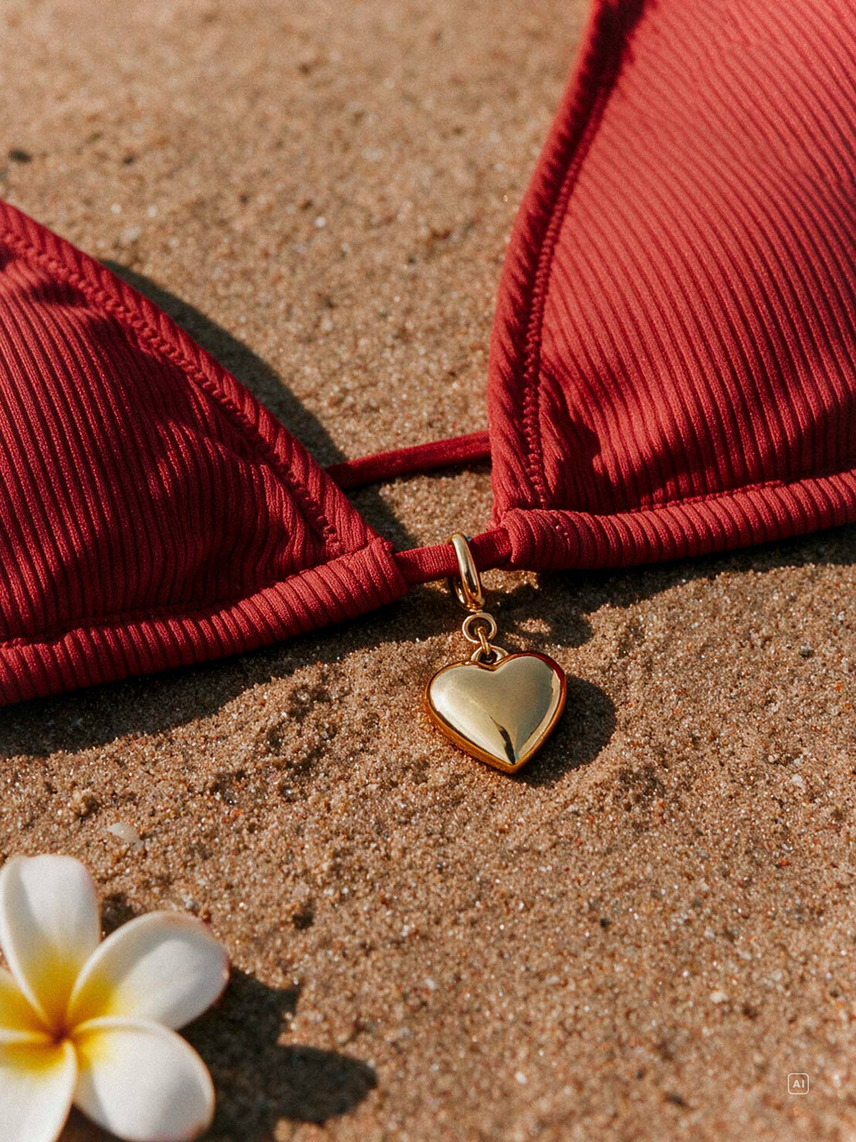 Goldfarbener Herz-Anhänger liegt auf Sand neben rotem Bikini-Oberteil und weißer Blüte.