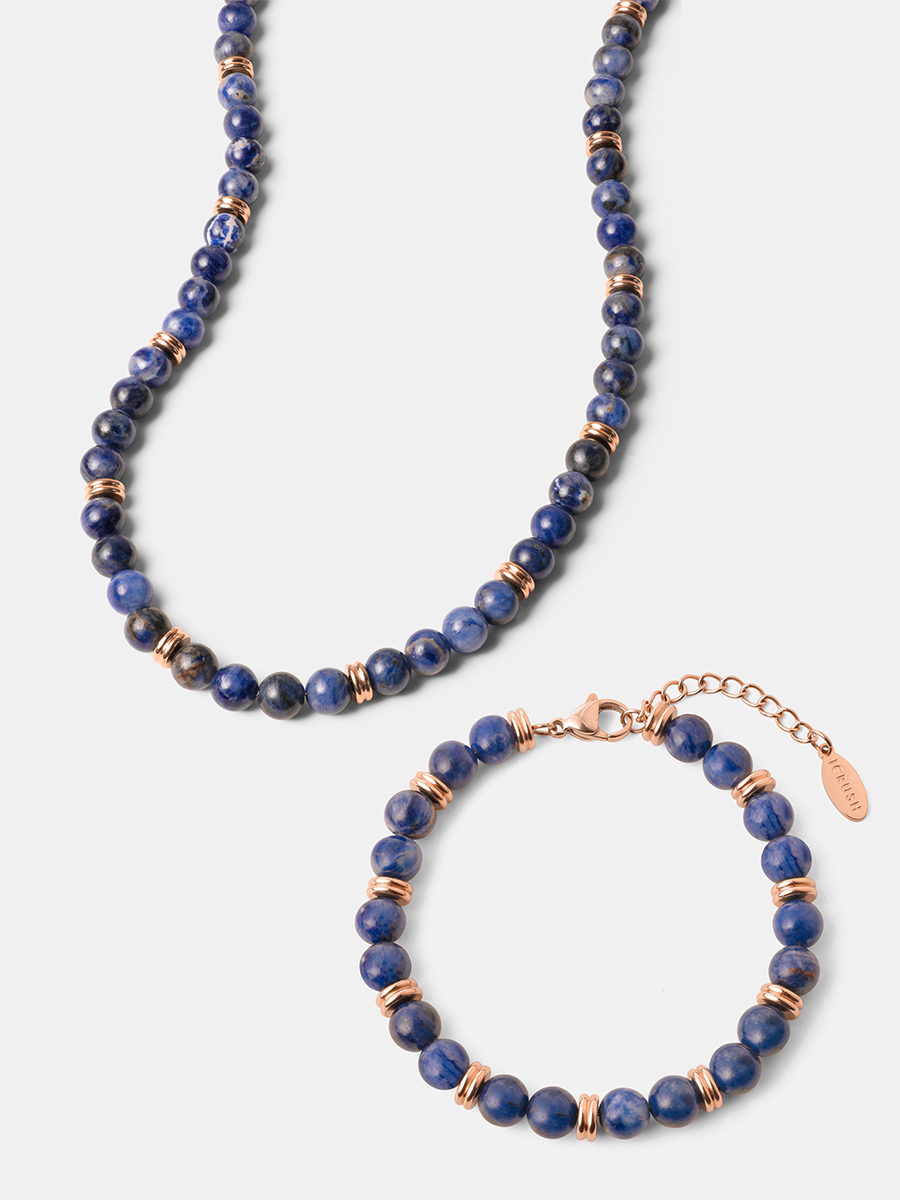 Dunkelblaue Perlenkette neben Armband mit roséfarbenen Metallringen, Verschluss und kleinem Anhänger auf grauem Hintergrund.