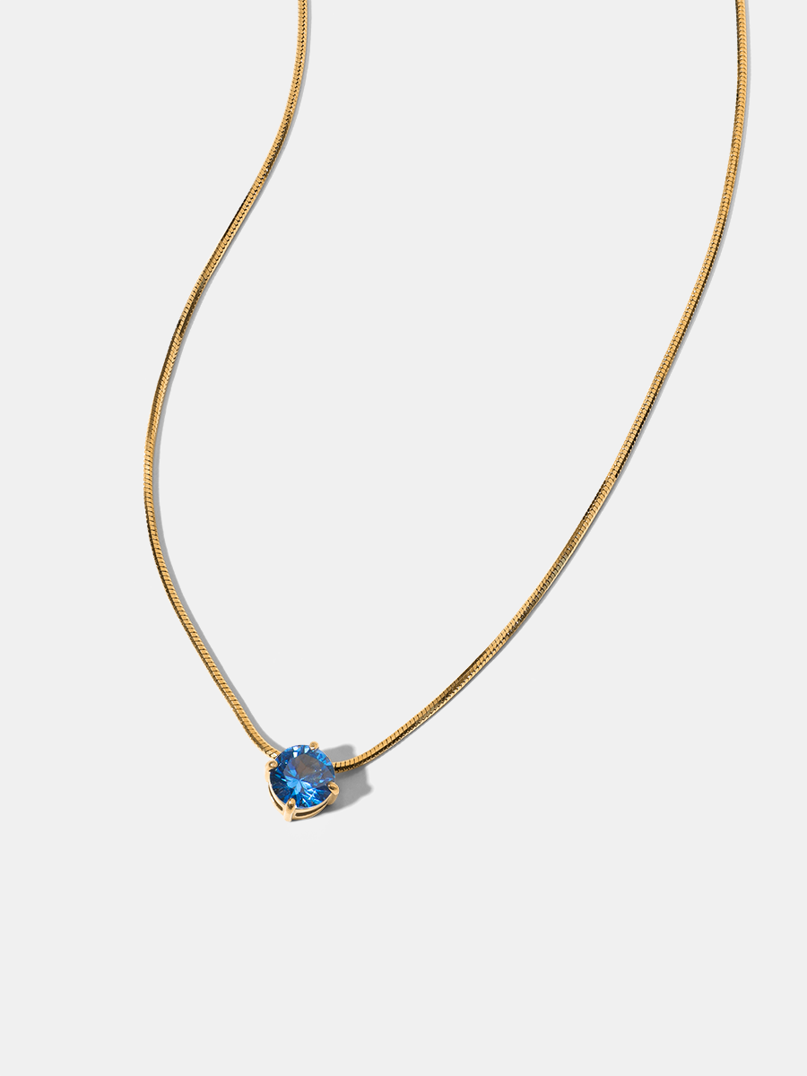 Goldfarbenes Collier mit blauem Facettstein und feiner Kette, präsentiert vor hellgrauem Hintergrund.