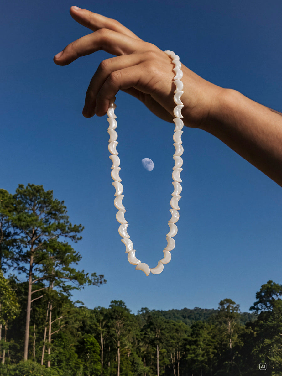 Hand hält goldfarbene Lunar Strand Kette mit weißen, geschwungenen Gliedern vor blauem Himmel, Wald und Mond.