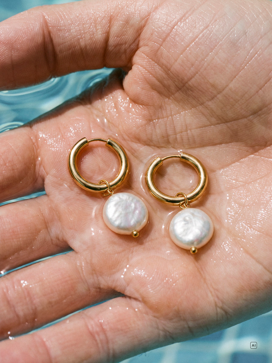 Hand hält zwei goldene Creolen mit weißen Perlen, Wasser und nasse Haut sichtbar.