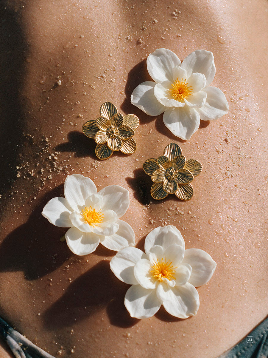 Zwei goldfarbene Blüten-Ohrringe liegen auf gebräunter Haut neben weißen Blüten mit gelber Mitte.