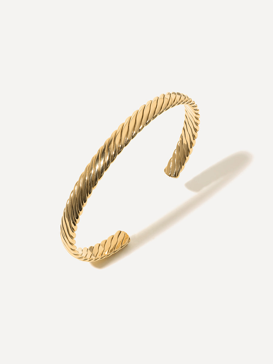 Goldener Armreif mit Seiltextur und offener Form liegt auf hellem Hintergrund, Schatten fällt rechts.