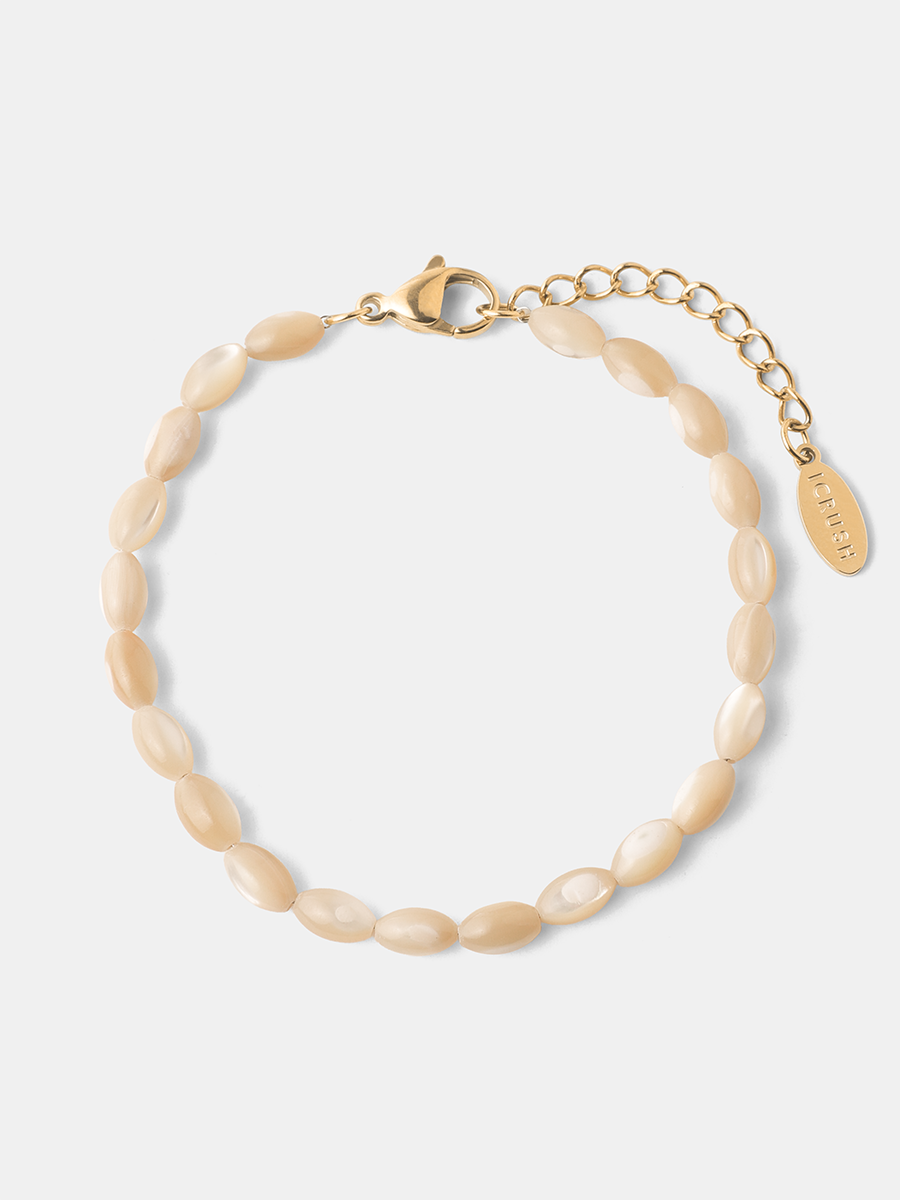 Goldfarbenes Muschelperlenarmband mit Karabinerhaken, Verlängerungskette und ovalem Markenanhänger auf neutralem Grund.