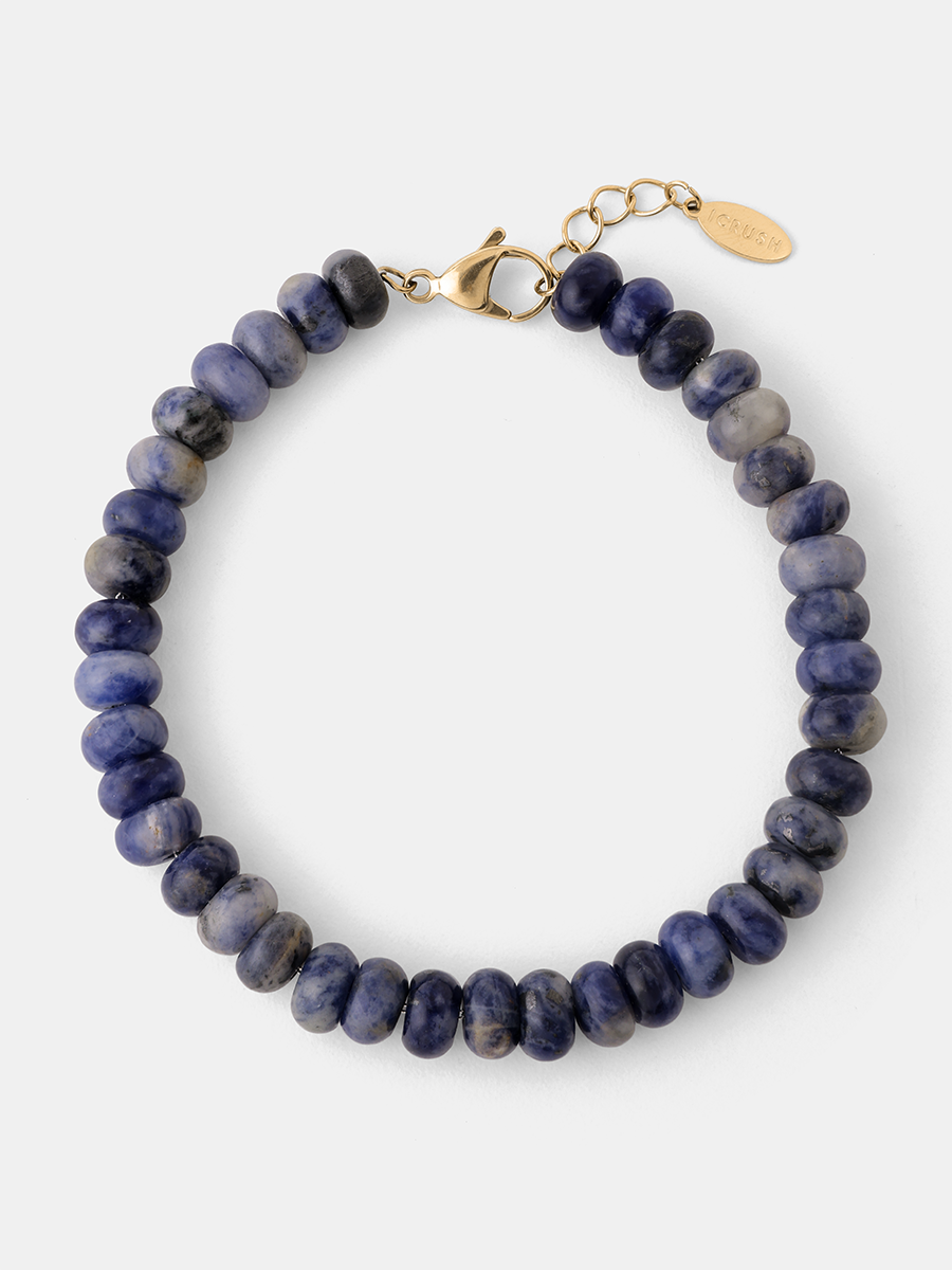 Rundes Lapisarmband mit goldfarbenem Karabiner und Markenplättchen, freigestellt.