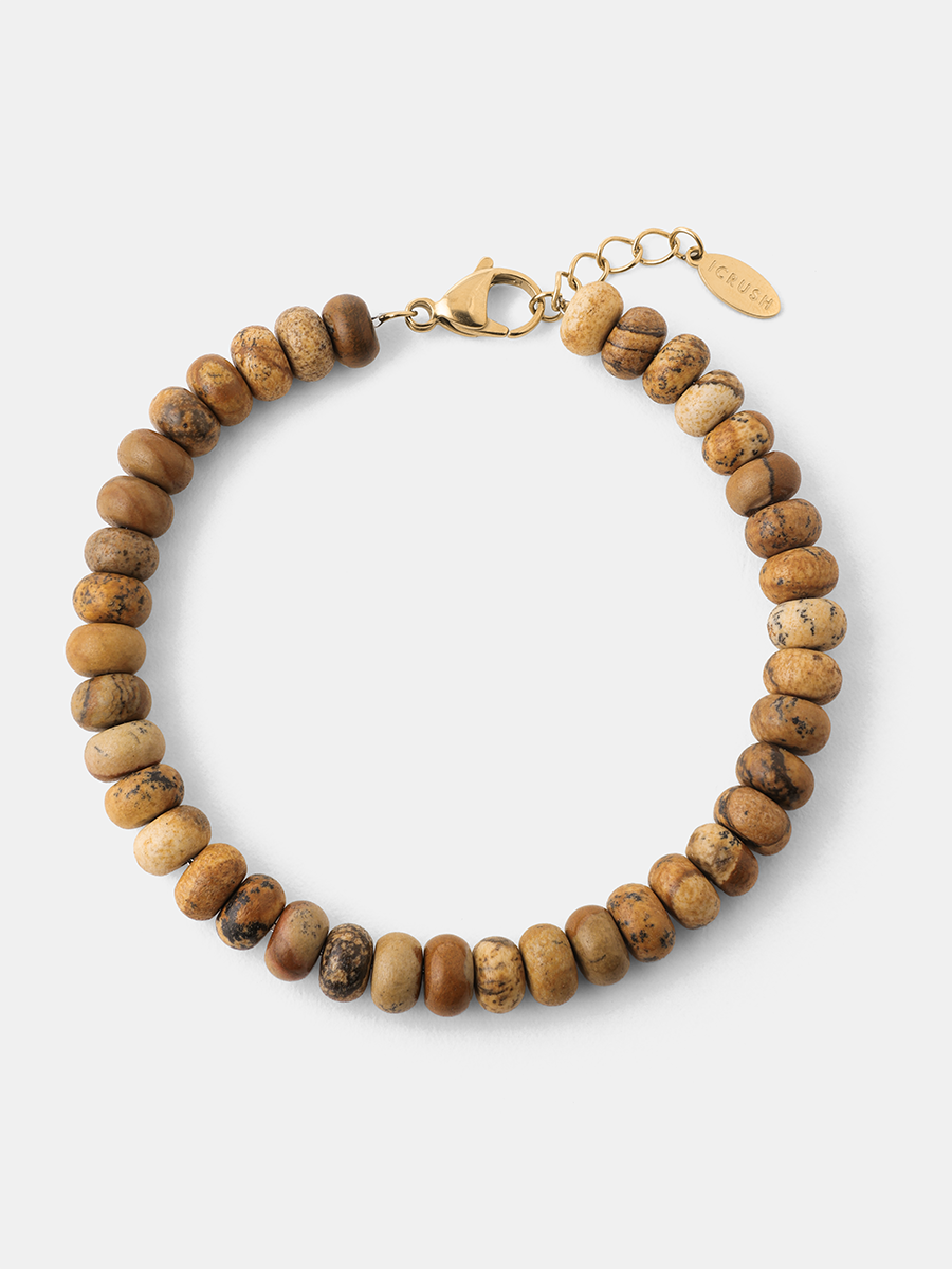 Goldfarbenes Karabinerarmband mit braunen, runden Steinperlen, Verlängerung und ovalem Logoanhänger auf hellem Untergrund.
