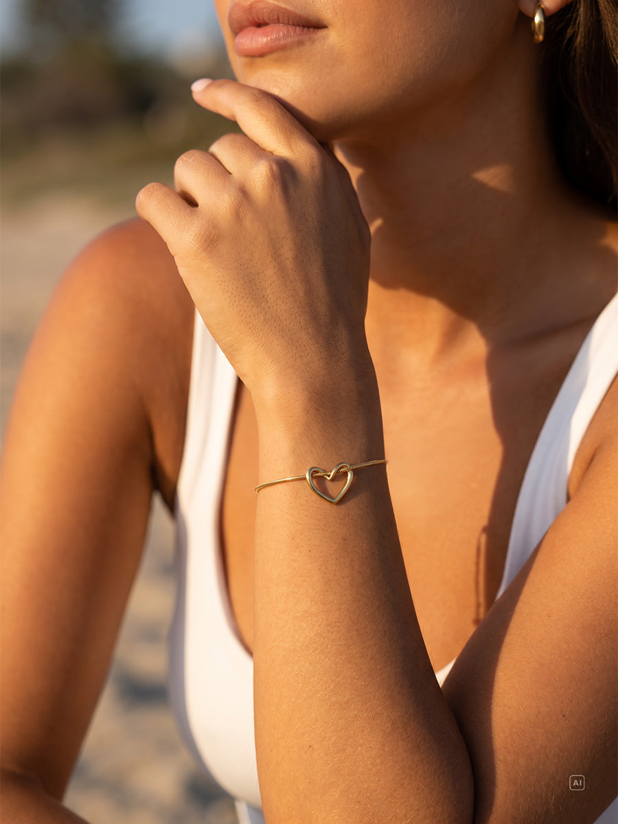 Frau mit weißem Top trägt goldenen Handgelenkschmuck mit offenem Herzanhänger, dahinter verschwommener Strand.