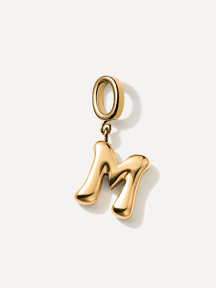 Goldglänzender Buchstaben-Charm M, kompakt und rundlich, mit Aufhängering, vor einfarbig hellem Grund.