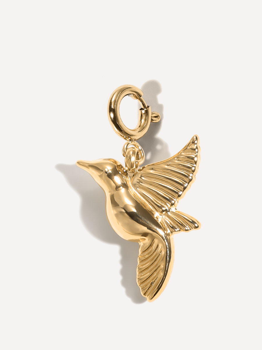 Goldener Kolibri-Charm mit ovaler Öse, detaillierten Flügeln und weichem Schatten vor hellem Hintergrund.