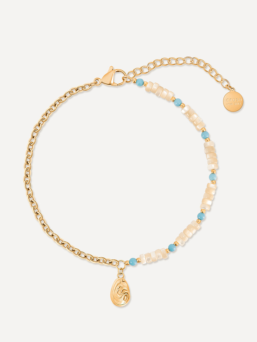 Goldfarbenes Fußkettchen flach ausgelegt, mit Gliederkette, beigen Perlen, türkisfarbenen Steinchen und ovalem Anhänger auf hellem Untergrund.