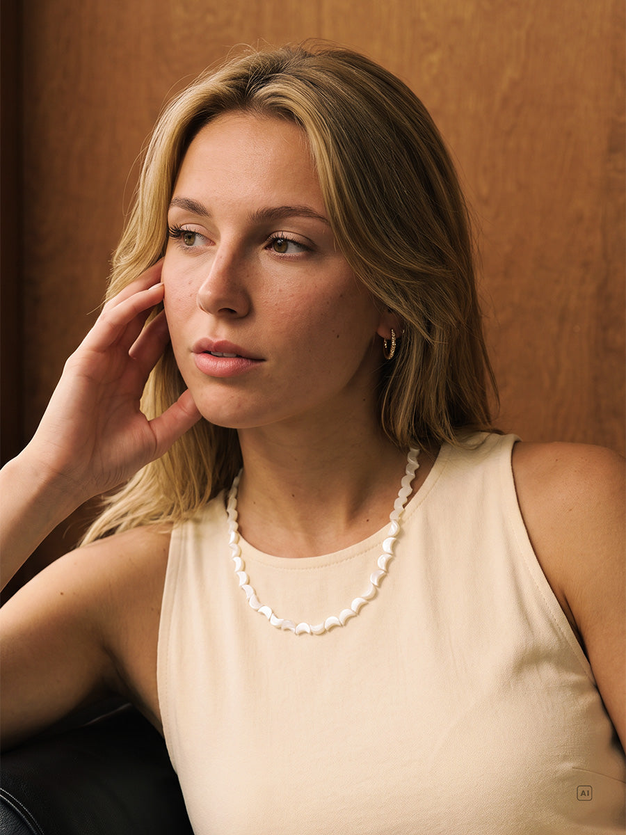 Frau mit beigem Top und goldfarbener Lunar Strand Kette blickt nachdenklich zur Seite, Holzwand im Hintergrund.