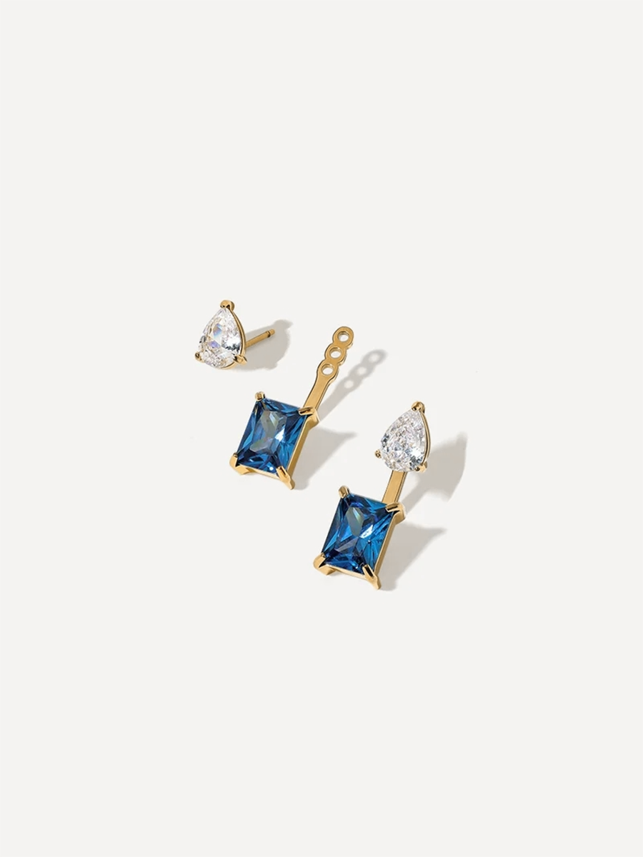 Goldene Ohrringe als Stecker mit blauem Viereck-Anhänger und Klarstein, liegend auf hellem Untergrund.